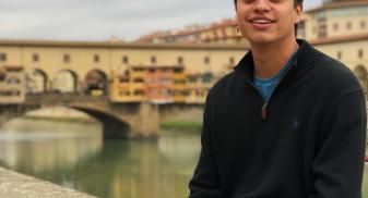 John is in a sweater and jeans sitting on a wall overlooking the river in Florence