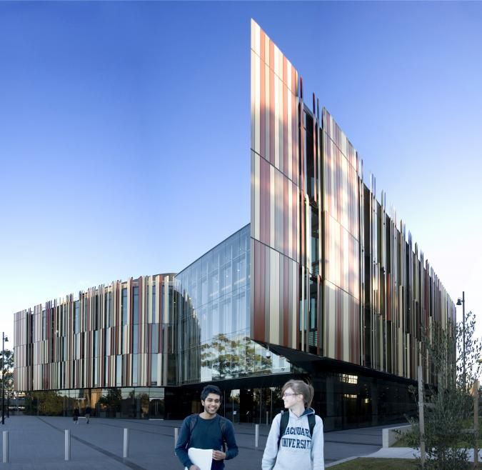 Two Macquarie students walk and laugh. Behind them is a large building with many glass panels of different reds and taupe. 