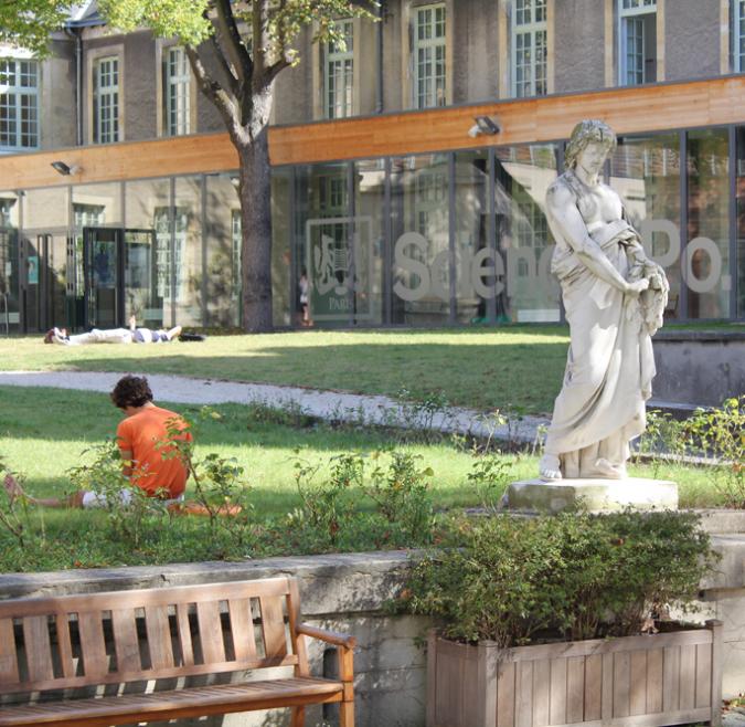 A marble statue sits before a Reims campus quad. Students are seen studying and lounging in the grass, and the Sciences Po logo is in the background on a window of the building