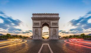 The Arc de Triomphe is in the center with the sun setting behind it and the headlights of cars stream by